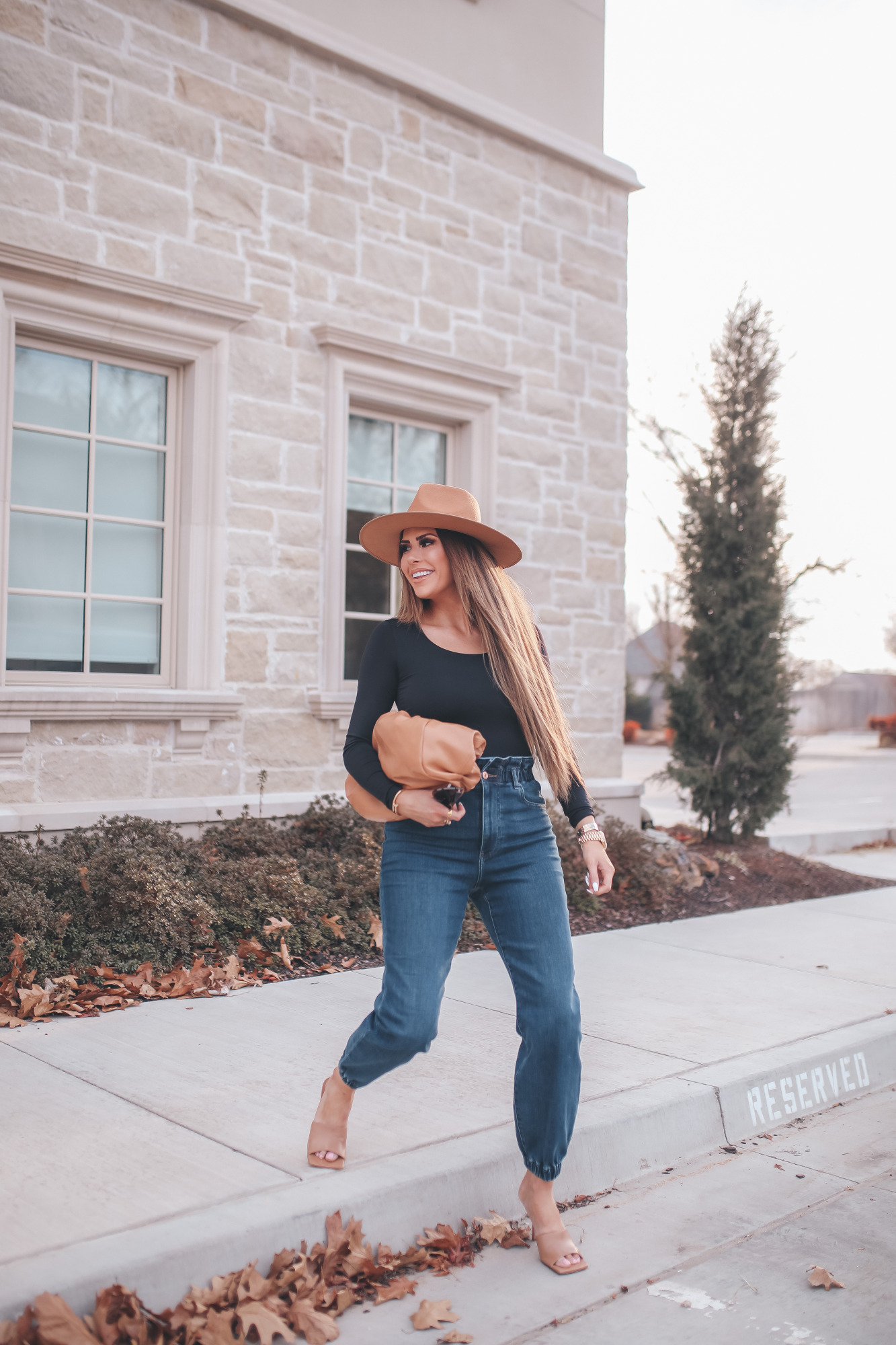 express spring fashion 2021, bottega clutch dupe, denim high waisted joggers, emily gemma15 |Body Contour by popular US fashion blog, The Sweetest Thing: image of a Emily Gemma wearing an Express black bodysuit, Express high waisted joggers, tan heel slide sandals, tan felt fedora, and holding a Bottega clutch dupe. 