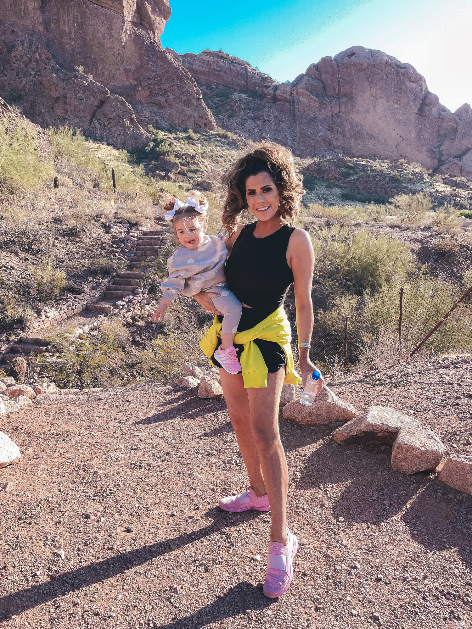 free people shorts, best workout gear spring 2021, camelback mountain, alo yoga top, emily gemma12 |Hiking Outfits by popular US fashion blog, The Sweetest Thing: image of Emily Gemma wearing a Alo yoga top, Nike half zipper pullover sweatshirt, Free People shorts, APL sneakers and Amazon sunglasses while holding her daughter Sophie who's wearing a grey and white polka dot lounge set, sneakers, and matching white hair bows. 