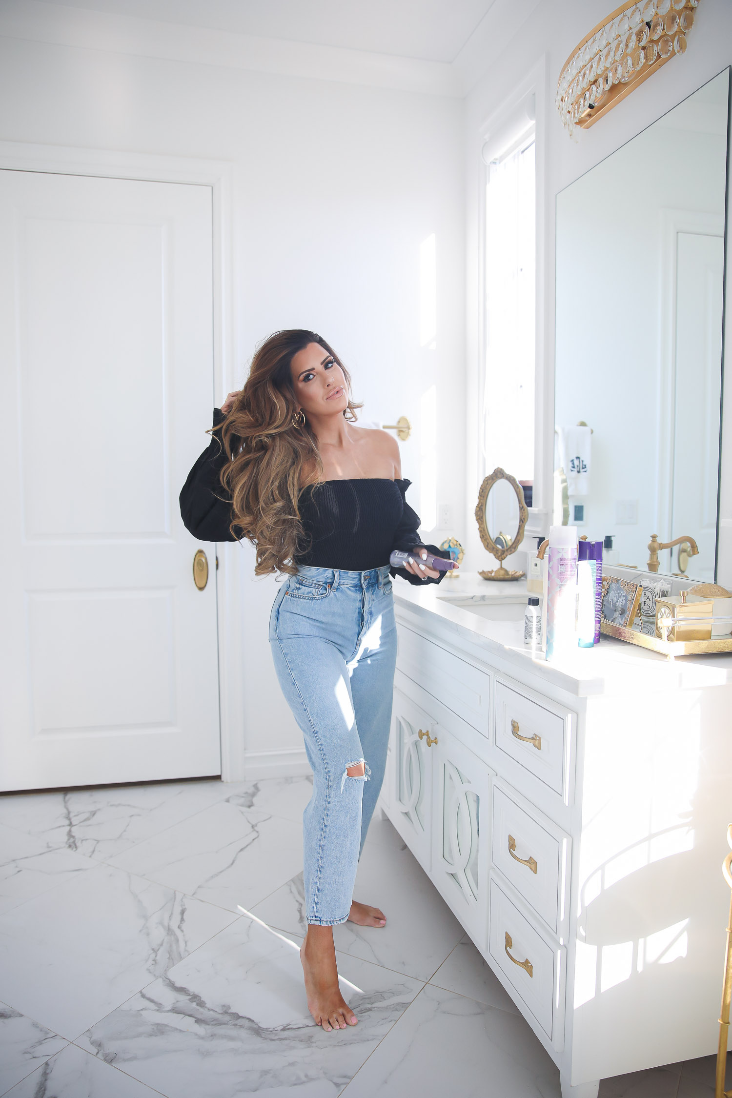 hair dot com, best hair products for long hair and extensions, Purology shampoo and conditioner, emily gemma, the sweetest thing blog | Hair Products by popular US beauty blog, The Sweetest Thing: image of Emily Gemma standing in her bathroom and wearing a black off the shoulder top, high waisted light denim jeans, and holding a bottle of Pureology hair product in her hand. 