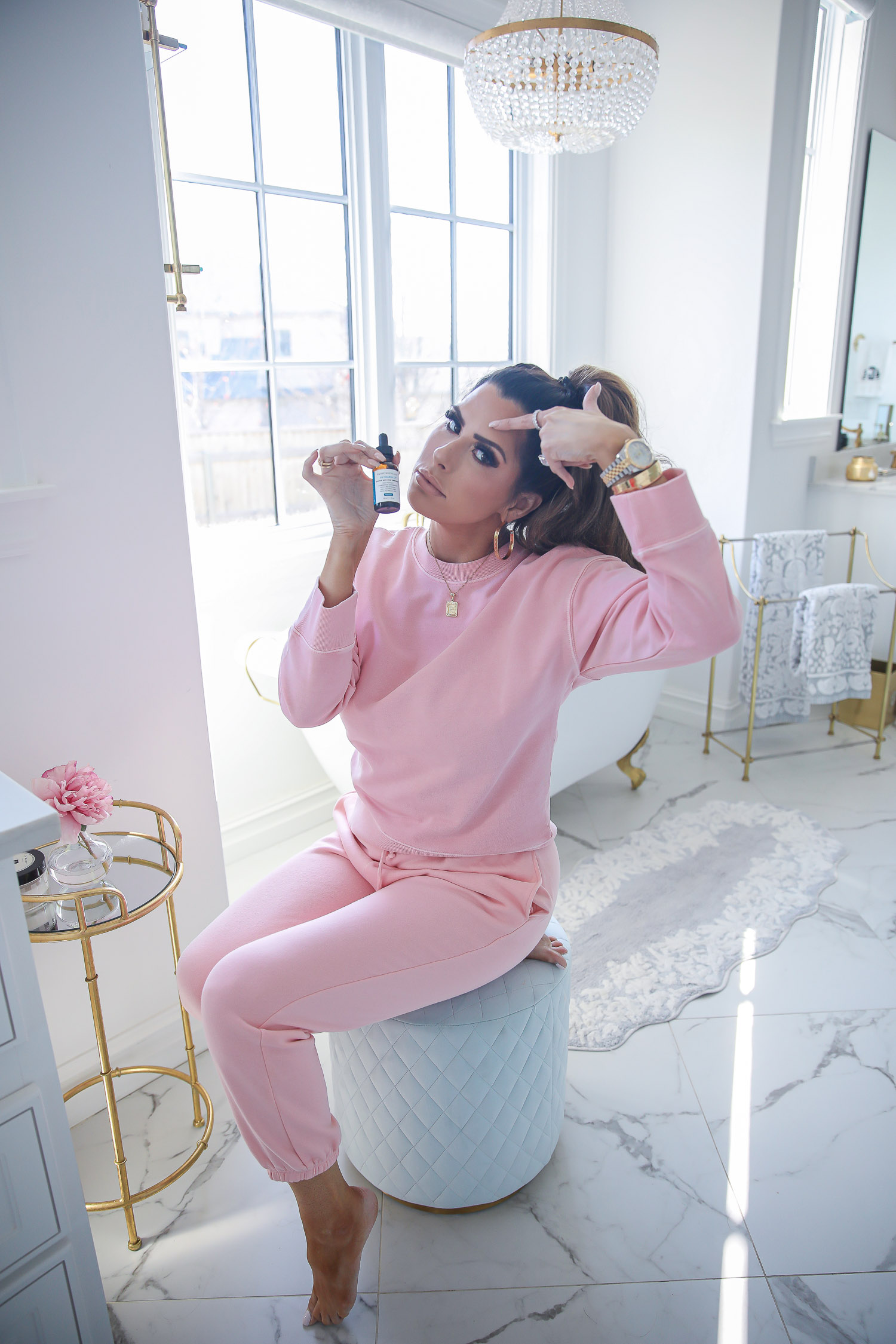 Skinceuticals CE Ferulic SilyMarin Phloretin CF, best Vitamim C serum, CE Ferulic vs Phloretin CF, why do you need Vitamin C, emily gemma skincare |Vitamin C Skincare by popular beauty blog, The Sweetest Thing: image of Emily Gemma sitting on a blue tuft stool in her bathroom and wearing a pink sweatsuit while holding a bottle SkinCeuticals skin care product. 
