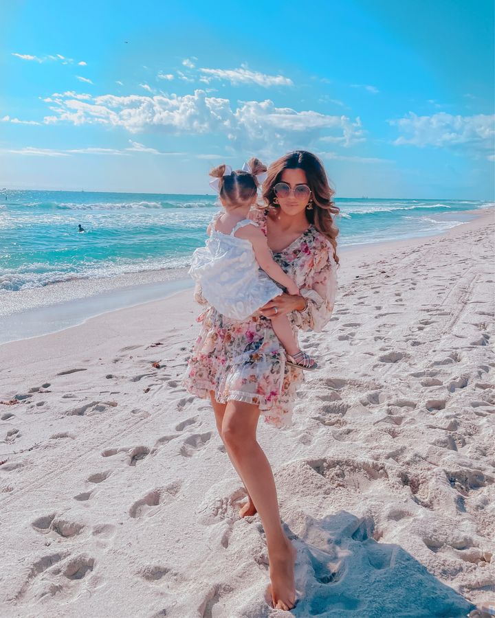 Floral Zimmermann Dress, Emily Gemma, Family Beach Photo, Emily Gemma, Dior Sunglasses, Chanel Earrings, Emily Ann Gemma | April Instagram Recap by popular US fashion blog, The Sweetest Thing: image of Emily Gemma holding her daughter Sophie on the beach and wearing a white floral print dress, gold Chanel earrings and floral print mini dress with long sleeves.