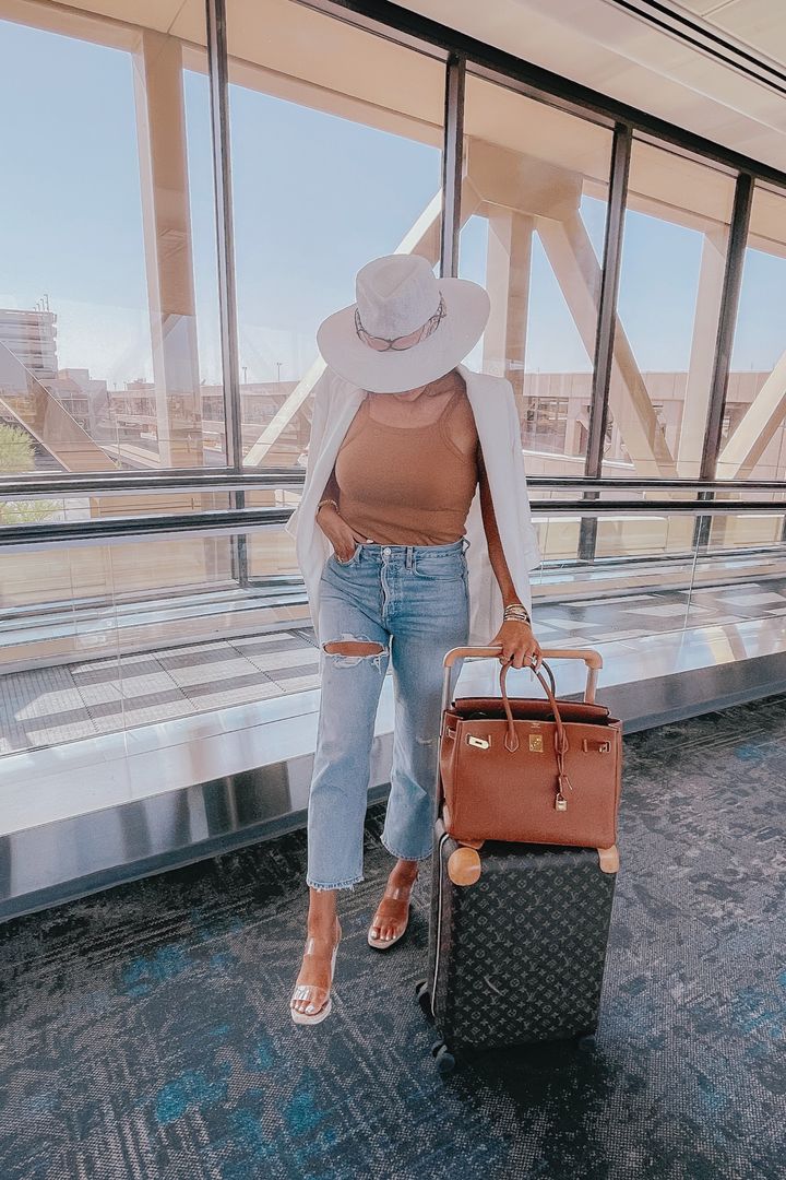 Emily Ann Gemma, Emily Gemma airport outfit, airport outfit ideas, linen cotton white blazer, hermes birkin, togo gold, white straw hat, Louis Vuitton carry on suitcase, agolde high waisted denim, clear espadrille wedge heels, Steve Madden wedges, travel outfit | Instagram Recap by popular US life and style blog, The Sweetest Thing: image of Emily Gemma wearing a white fedora, white linen blazer, tan tank top, light wash high waisted jeans, and clear strap sandals while pulling a Louis Vuitton rolling suitcase with a brown designer bag resting on top. 