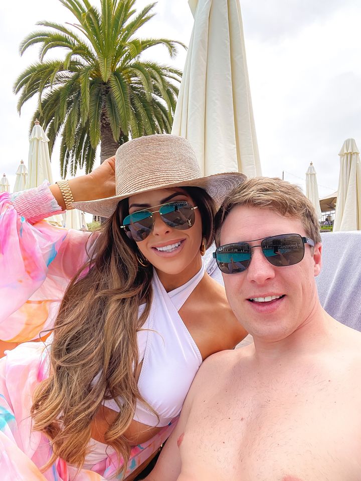 Couple hanging out by the pool, Emily Gemma, John Gemma, Straw beach hat, white swimsuit, white bikini top, saint laurent sunglasses, colorful swim cover up, laguna beach | Instagram Recap by popular US life and style blog, The Sweetest Thing: image of Emily Gemma sitting on a lounge chair at the beach and wearing aviator sunglasses, straw hat, white two piece swimsuit and pink tropical print chiffon coverup. 