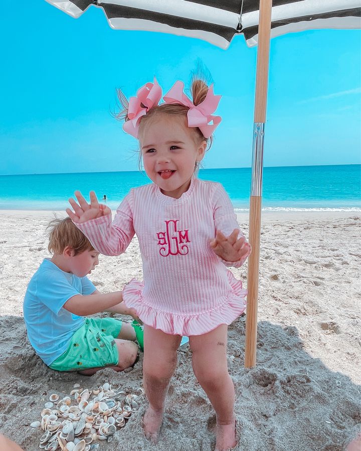 Sophie Gemma, Luke Gemma, Swim wear, Toddler Swim Suits, Monogrammed Swimsuit, Beachwear, Emily Gemma | April Instagram Recap by popular US fashion blog, The Sweetest Thing: image of a young boy and girl playing in the sand under a black and white stripe shad umbrella and wearing pink hair bows, pink and white stripe monogram Smock Auctions swimsuit, white rash guard, and green swim shorts. 