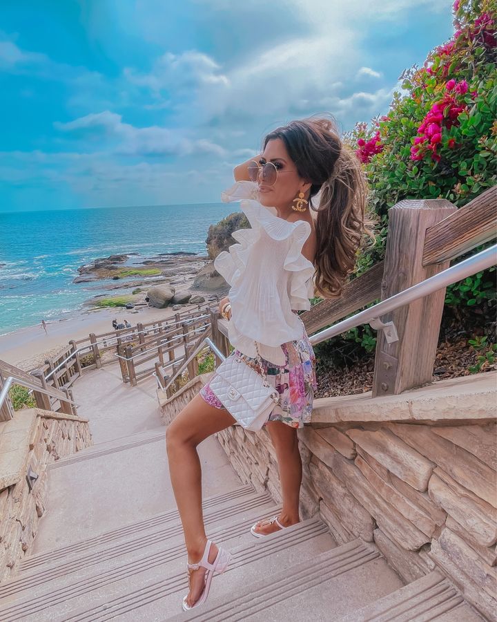 Zimmermann top, Cettire, Chanel pink sandals, Emily Ann Gemma, Ruffle long sleeve crop top, dior round sunglasses, high ponytail, laguna montage , chanel earrings | Instagram Recap by popular US life and style blog, The Sweetest Thing: image of Emily Gemma standing on some cement stairs near the ocean and wearing a white long sleeve crop top, gold Chanel statement earrings, round sunglasses, floral print shorts, white sandals, and holding a white quilted purse. 