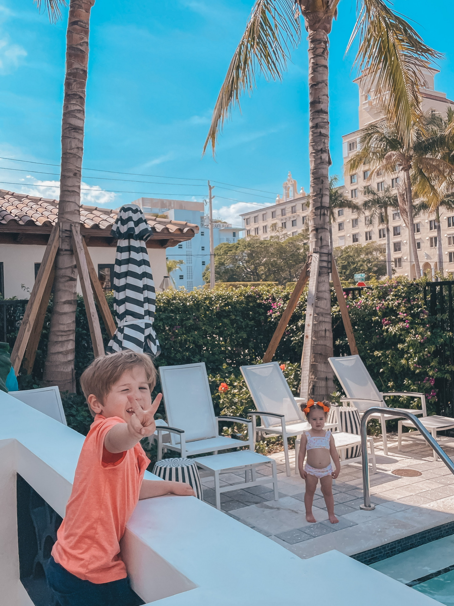 High Waisted Bikini by popular US fashion blog, The Sweetest Thing: image of a young boy wearing a orange polo and blue pull-on shorts and young girl wearing orange hair bows and two piece swimsuit standing by a swimming pool and poolside white lounge chairs. 