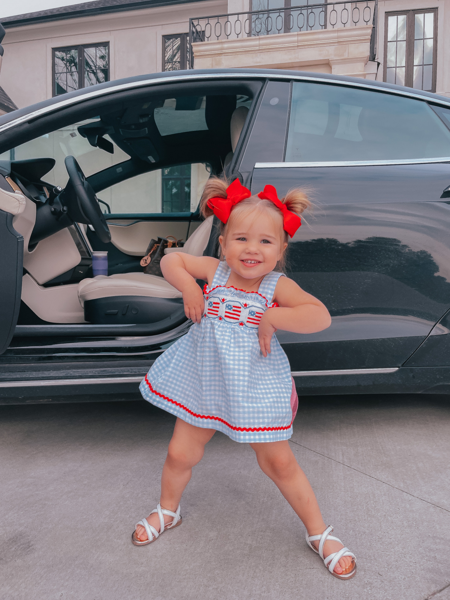 Sophia Gemma, American flag outfit for little girl, Fourth of July little girl outfit, blue and white checked bloomer set, red white and blue outfit ideas for toddlers, red hair bows for little girl. | Instagram Recap by popular US life and style blog, The Sweetest Thing: image of a little girl standing in front of a black car and wearing a smocked auctions 4th of July dress, two red bows and white strap sandals. 