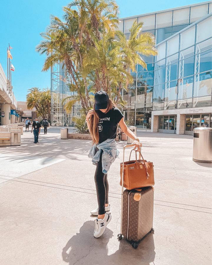 Emily Gemma airport travel outfit, airport outfit ideas, travel outfit ideas, Balmain t shirt, jean jacket, best leggings to wear to the airport, travel outfit ideas 2021, hermes birkin, Louis Vuitton carry on suitcase, black baseball cap | Instagram Recap by popular US life and style blog, The Sweetest Thing: image of Emily Gemma standing outside and wearing a black ball cap, black leggings, Chanel platform sneakers, black t-shirt, and light denim jacket tied around her waist and standing next to a Louis Vuitton rolling suitcase with a brown designer bag resting on top. 
