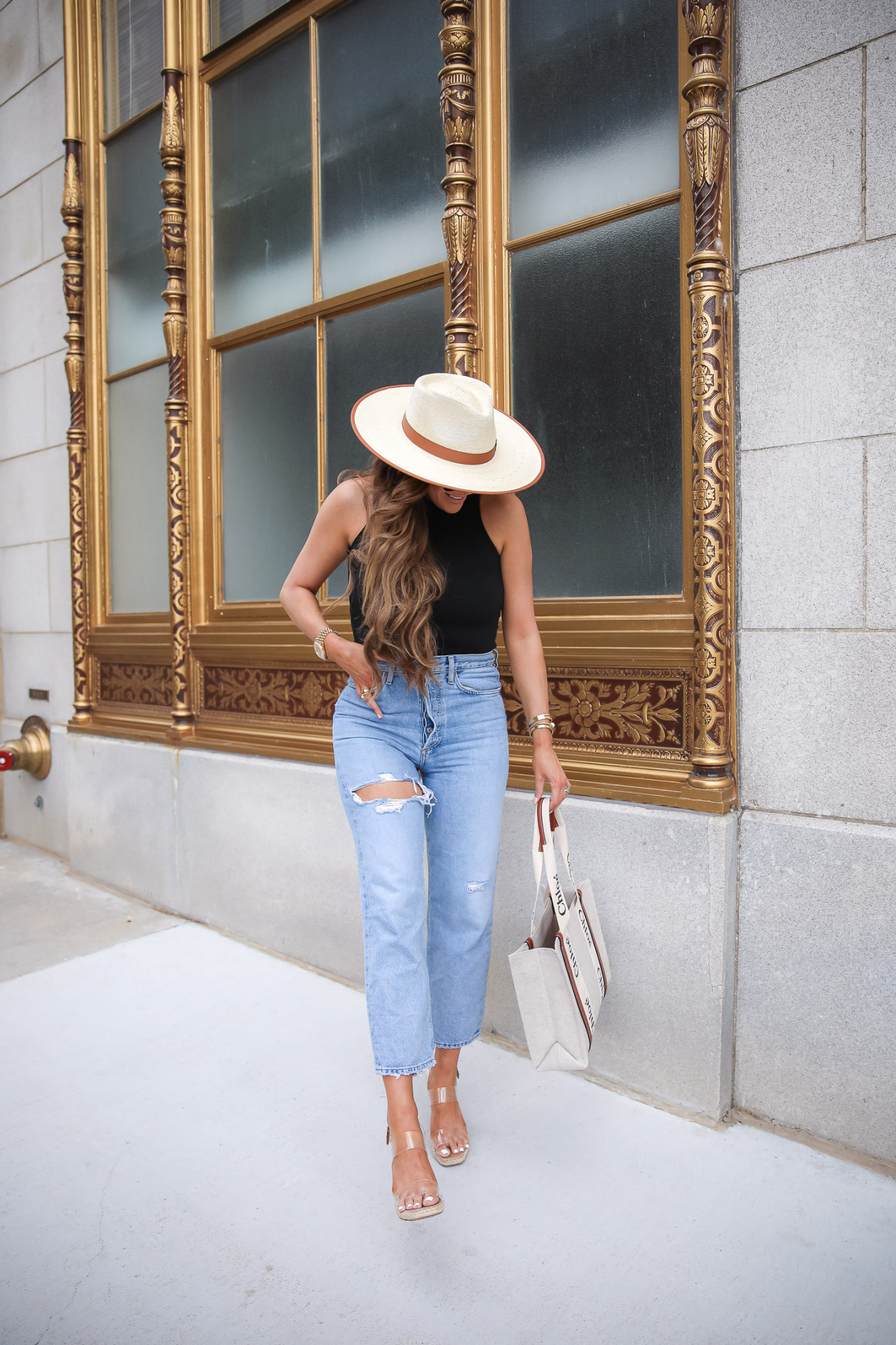 Summer Outfit by popular US fashion blog, The Sweetest Thing: image of Emily Gemma standing outside in front of a building with gold framed windows and wearing a cream fedora hat, BP mock neck tank, Agolde jeans, Steve Madden clear strap espadrilles, and holding a Chloe tote bag. 