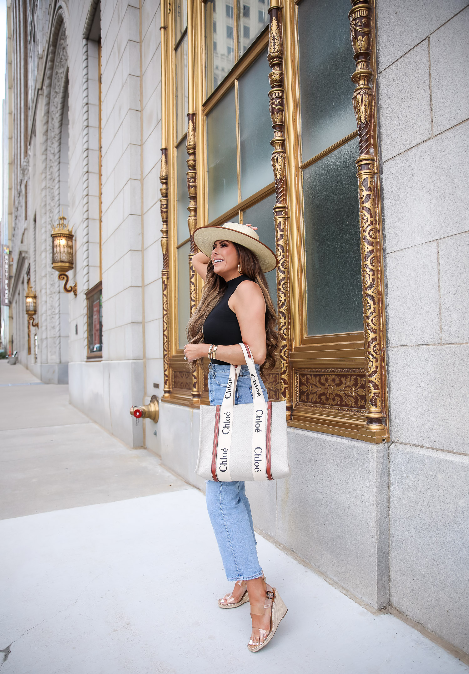 Summer Outfit by popular US fashion blog, The Sweetest Thing: image of Emily Gemma standing outside in front of a building with gold framed windows and wearing a cream fedora hat, BP mock neck tank, Agolde jeans, Steve Madden clear strap espadrilles, and holding a Chloe tote bag. 