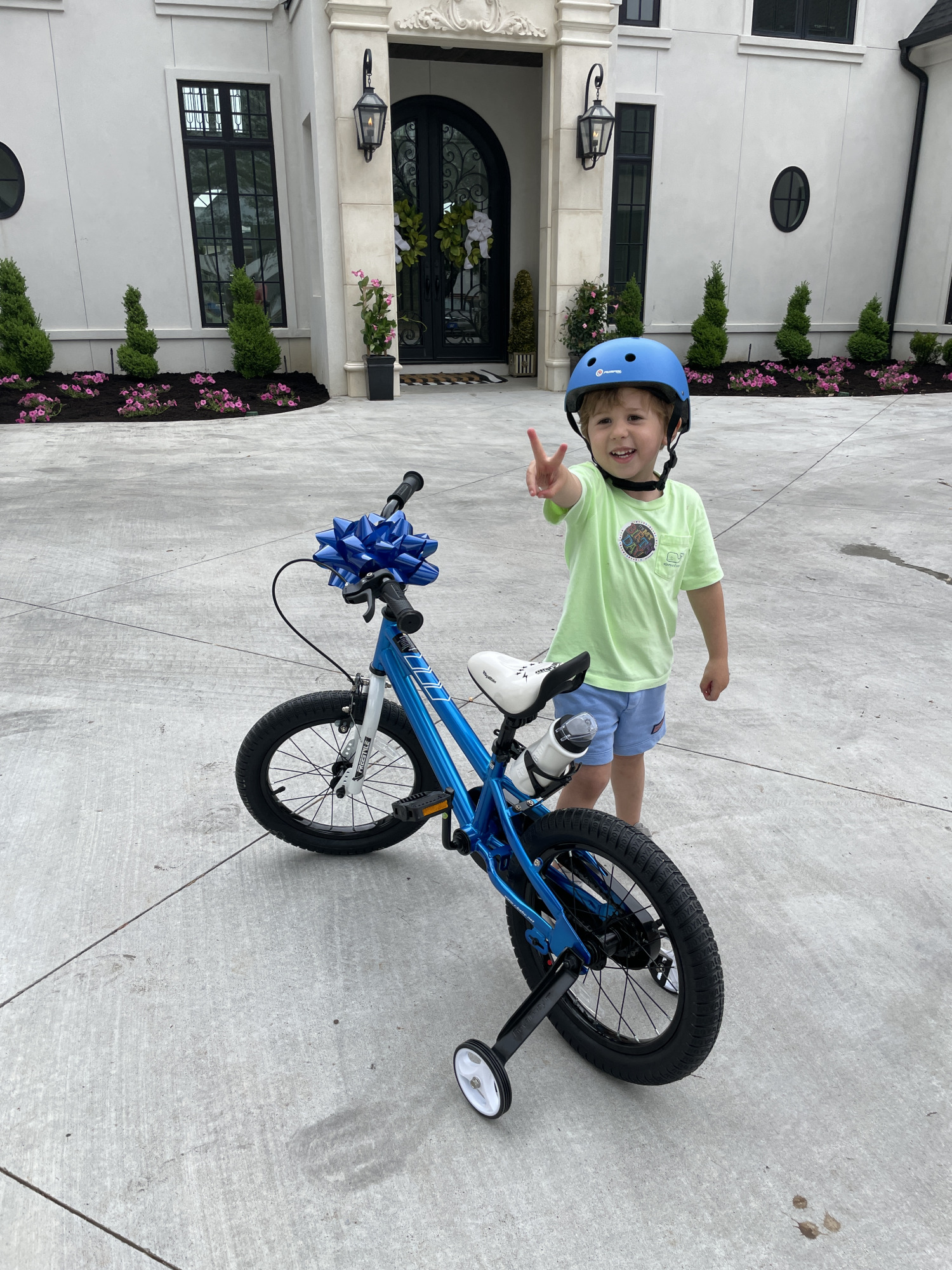 little boy gift ideas, bikes for little boys, bikes with training wheels, Luke gemma | Instagram Recap by popular US life and style blog, The Sweetest Thing: image of a little boy standing next to a blue bike with training wheels and holding up a the peace sign with his fingers and wearing a blue bike helmet, neon green shirt, and blue shorts. 