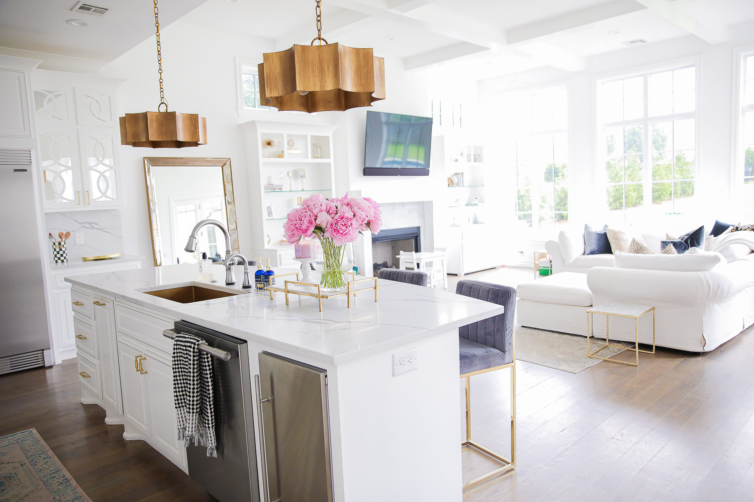 Pinterest white kitchen quartz, ilv majestic stove, Emily Gemma kitchen, gold kitchen sink, Emily Gemma kitchen, grey and gold barstools | Spring Kitchen by popular US life and style blog: image of a kitchen with white cabinets, wood flooring, wooden light pendants, marble counter tops, vase of pink peonies, grey velvet and gold metal bar stools and light pink and blue runner rug next to a living room with a white sectional couch, white marble and gold side table, fireplace, white shelving, and flat screen t.v.