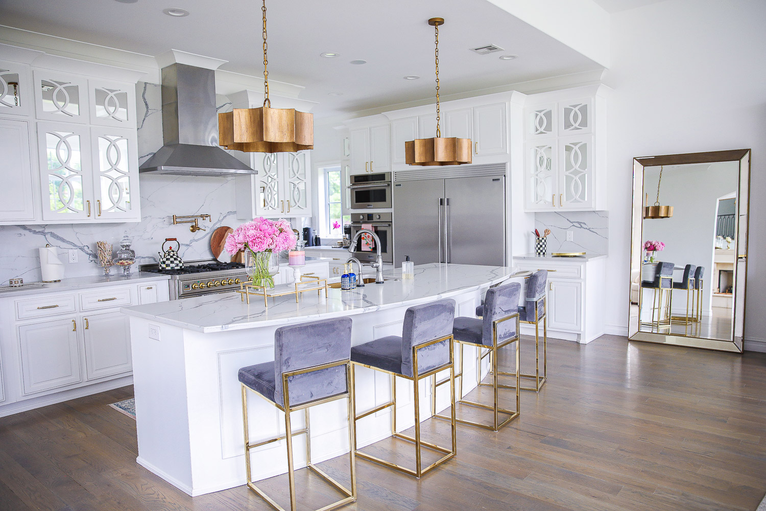 Spring Kitchen by popular US life and style blog: image of a kitchen with white cabinets, wood flooring, wooden light pendants, marble counter tops, gold and grey velvet bar stools, and light pink and blue runner rug. 
