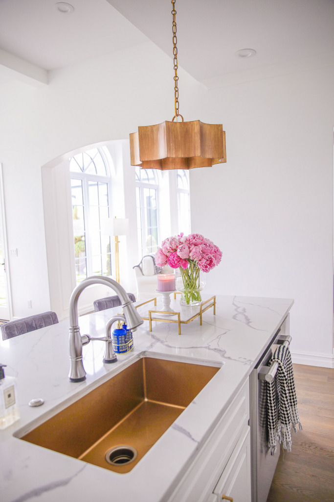gold kitchen sink, touch2o faucet, Pinterest white kitchen gold hardware, white kitchen inspiration, Emily Gemma kitchen | Spring Kitchen by popular US life and style blog: image of a kitchen with white cabinets, wood flooring, wooden light pendants, marble counter tops, grey velvet and gold metal barstools, vase of pink peonies, and light pink and blue runner rug. 