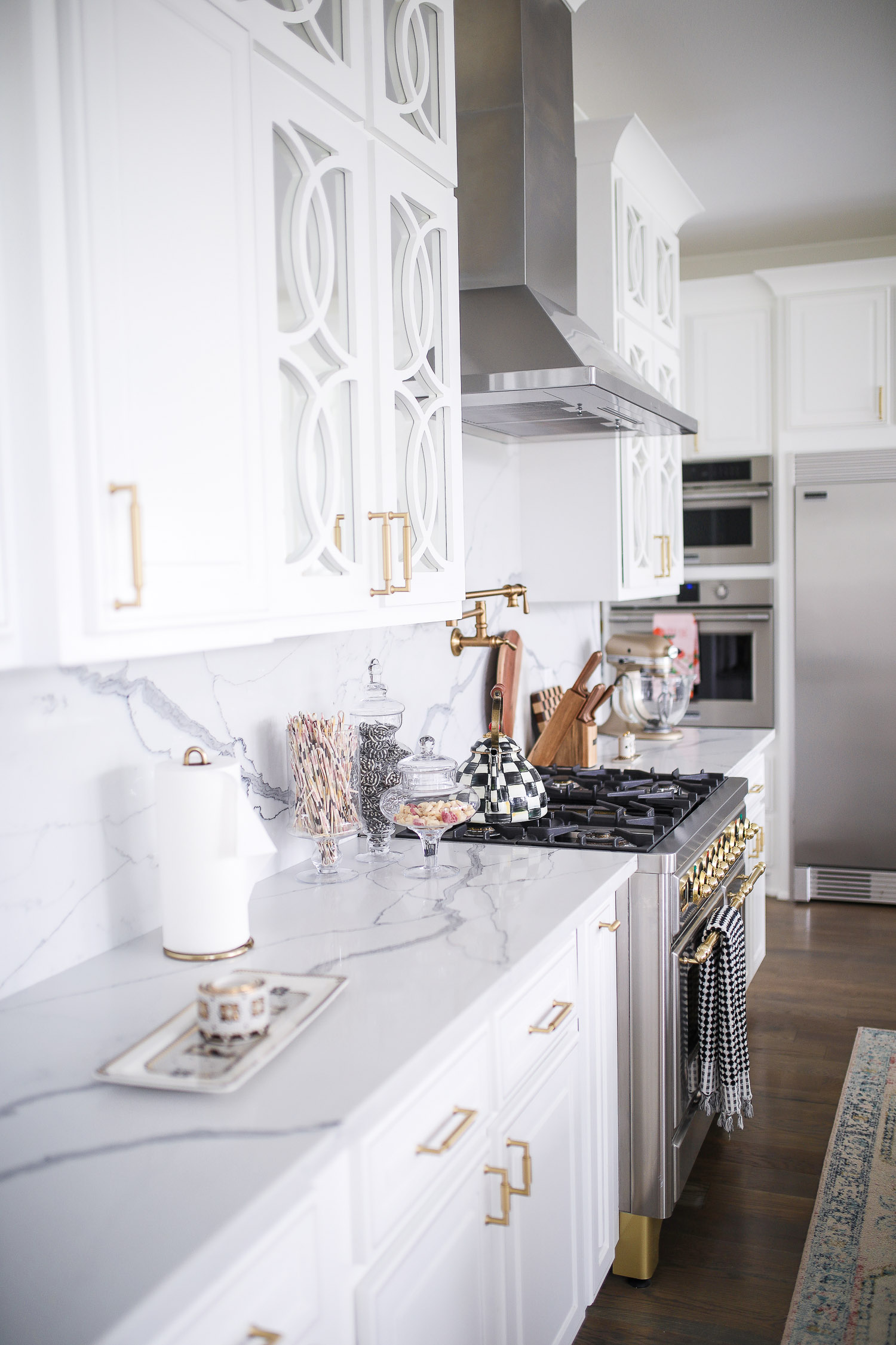 Pinterest white kitchen quartz, ilv majestic stove, Emily Gemma kitchen, gold kitchen sink, Emily Gemma kitchen, grey and gold barstools | Spring Kitchen by popular US life and style blog: image of a kitchen with white cabinets, wood flooring, wooden light pendants, marble counter tops, and light pink and blue runner rug. 