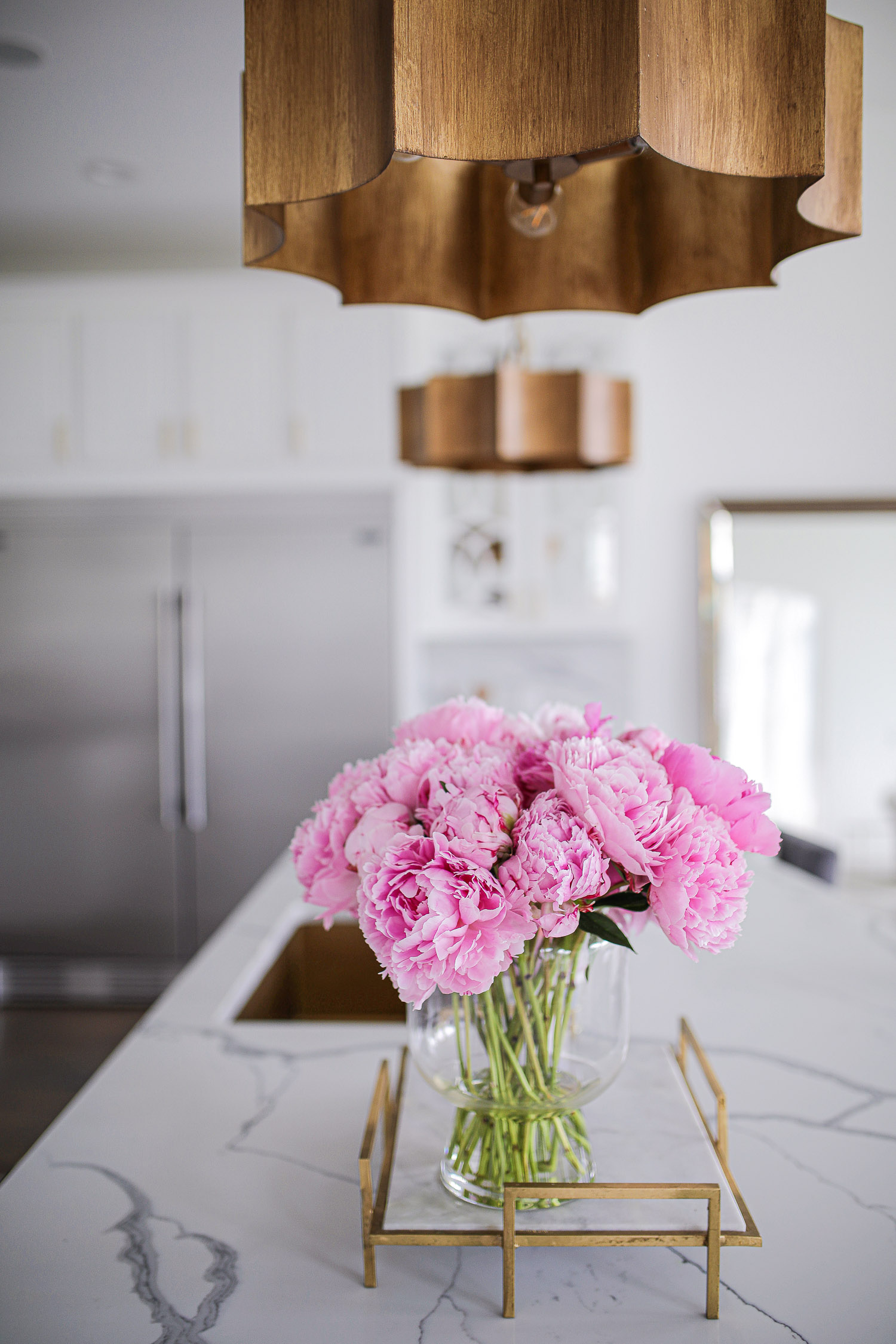 Pinterest white kitchen quartz, ilv majestic stove, Emily Gemma kitchen, gold kitchen sink, Emily Gemma kitchen, grey and gold barstools | Spring Kitchen by popular US life and style blog: image of a kitchen with white cabinets, wood flooring, wooden light pendants, marble counter tops, gold and marble tray with a  vase of pink peonies. 