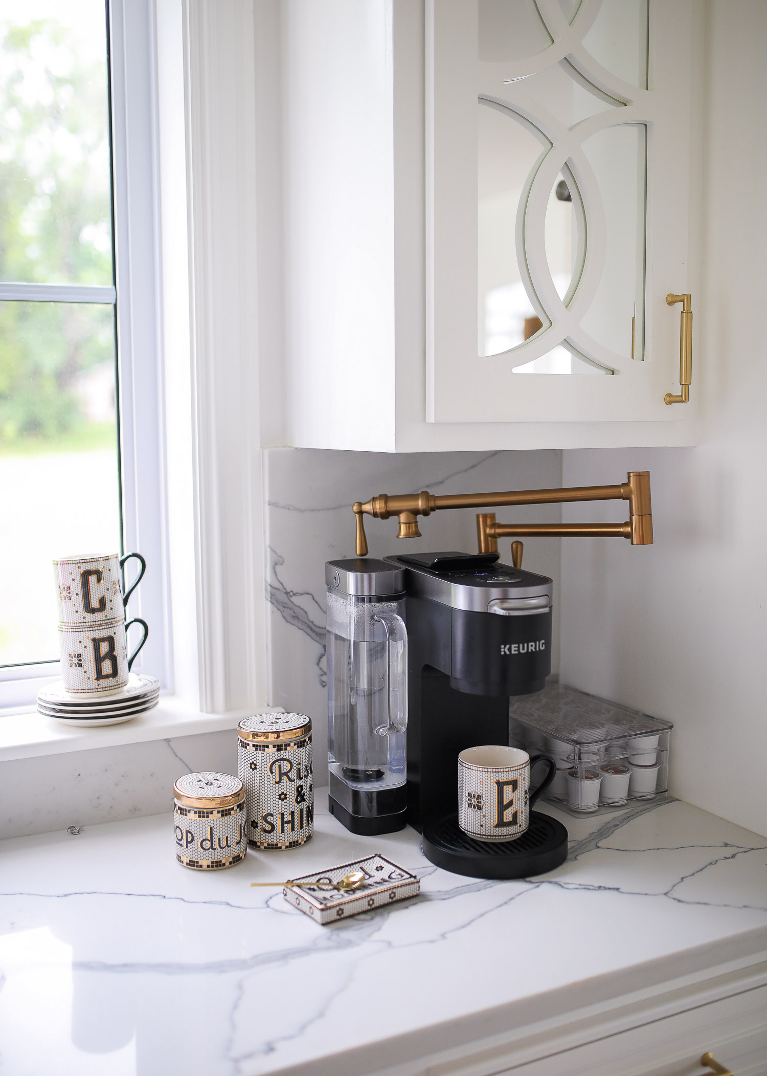 cute coffee bar, gold pot filler, pot filler for coffee maker, coffee bar ideas Pinterest, cute coffee bar Parisian dishes, Emily Gemma | Spring Kitchen by popular US life and style blog: image of a kitchen with white cabinets, marble counter tops, Anthropologie monogram mugs, and Keurig coffee station. 