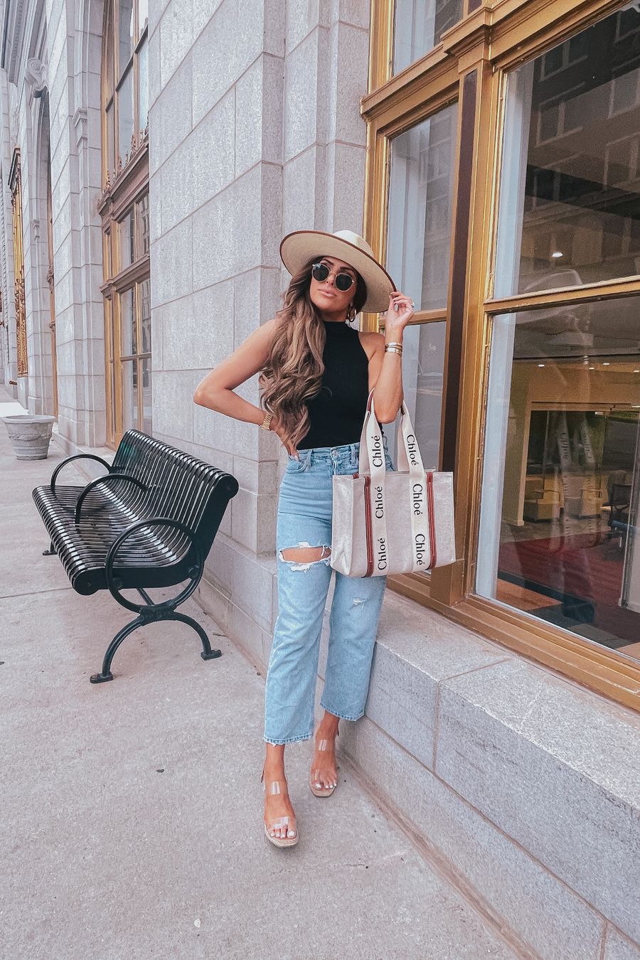 Summer Outfit by popular US fashion blog, The Sweetest Thing: image of Emily Gemma standing outside in front of a building with gold framed windows and wearing a cream fedora hat, BP mock neck tank, Agolde jeans, Steve Madden clear strap espadrilles, and holding a Chloe tote bag. 