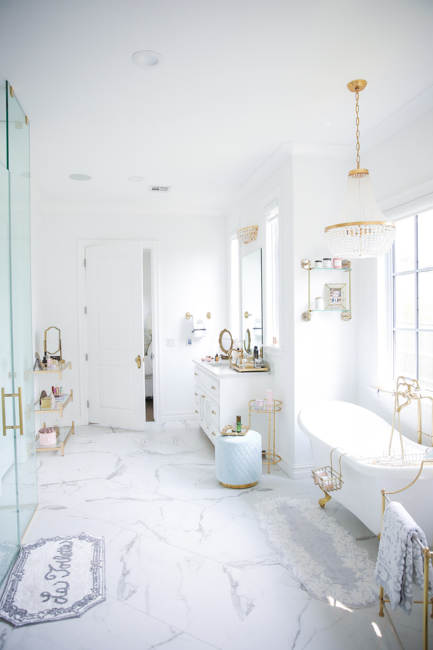 Emily gemma master bathroom, Nordstrom anniversary sale 2021 beauty must haves, anthropologie bathrom decor 2021, pinterest bathroom all white gold | Nordstrom Anniversary Sale 2021 by popular US life and style blog, The Sweetest Thing: image of a master bedroom with marble flooring, white and gold clawfoot tub, gold and crystal chandelier, gold metal towel stand and walk in shower. 
