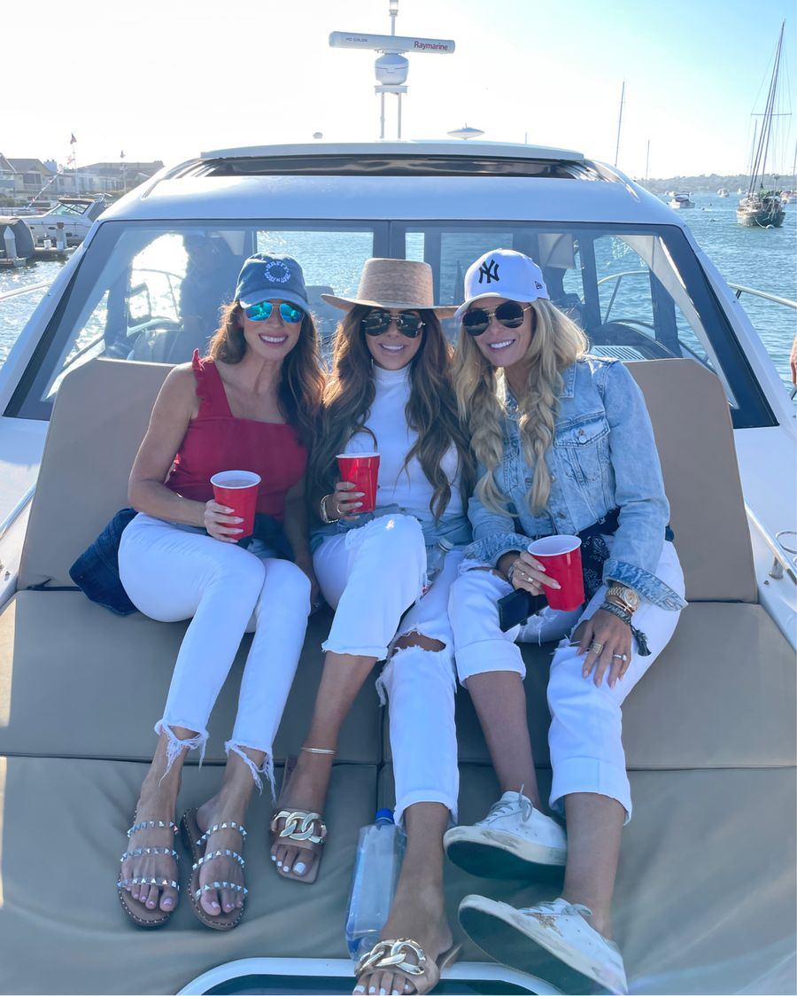 4th of July Outfits, White Denim, Best Sandals Summer 2021, Red White and Blue Outfit, What to Wear in Newport Beach, Boat Outfits, Emily Ann Gemma | Instagram Recap by popular US life and style blog, The Sweetest Thing: image of Emily Gemma sitting with two of her girlfriends on a boat and wearing a white mock neck shirt, Lack of Color boater hat, white distressed jeans, and tan and gold chain strap slide sandals. 