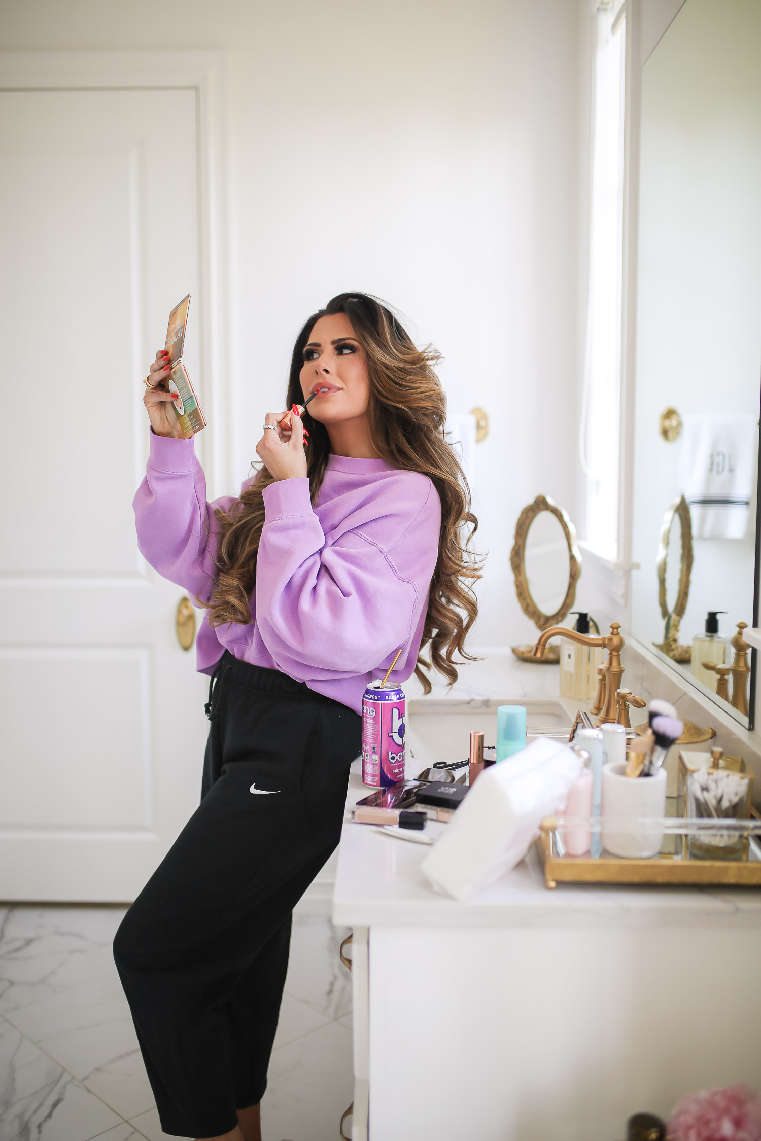 nordstrom beauty must haves fall 2021, sisley pore minimizer review, sisley blur powder review, sisley hyaluronic acid, emily gemma beauty must haves, hoola bronzer palette | Beauty Favorites by popular US beauty blog, The Sweetest Thing: image of Emily Gemma wearing a purple sweatshirt and black Nike jogger pants while leaning against her bathroom vanity and applying lipgloss while she looks in a handheld mirror. 
